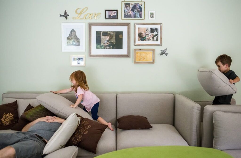 A little boy and girl hold play on the couch while their dad tries to sleep by documentary family photographer Polina Bulman.