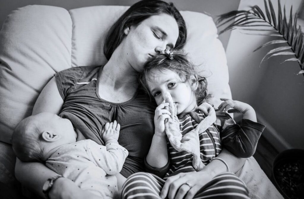 A mom snooze while her baby nurses and toddler chews a toy by documentary family photographer Polina Bulman.