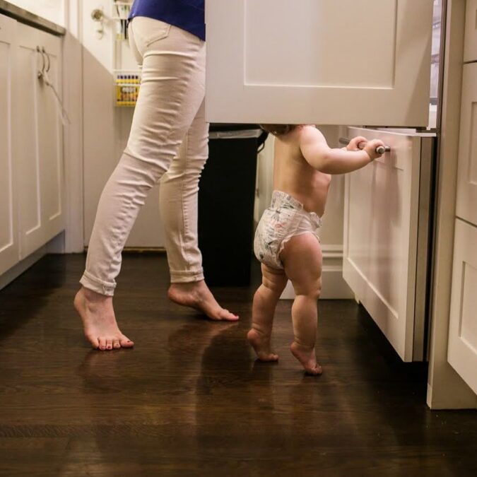 A mother and baby stand on tippy toes in the kitchen by documentary family photographer Polina Bulman.