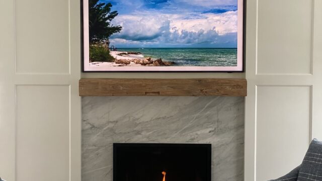 Cosy sitting room with a wall-mounted framed television above a lit fireplace and a coffee cup sitting on the coffee table JL Entertainment media installation
