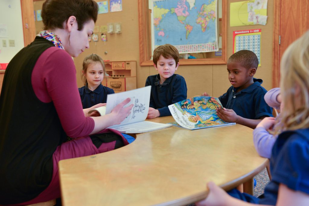 Children sitting at a table with their teacher about to begin their reading lesson.